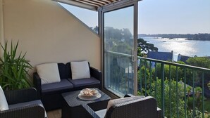 Ein Balkon mit Blick auf ein Gewässer, eine Sitzecke mit Sofa, ein Tisch mit einer Korb-Auswahl an Gebäck und ein gestreifter Sonnenschirm.