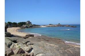 A coastal landscape with a large rock formation in the foreground, a sandy beach, and a building on a rocky outcrop in the background.