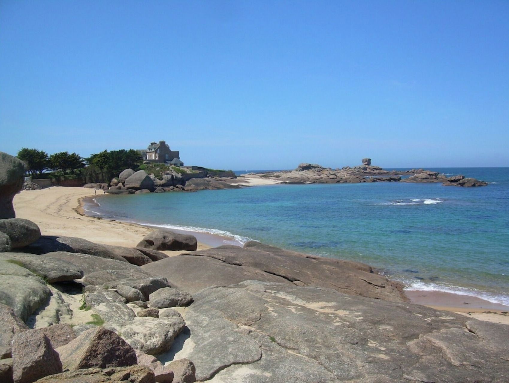 Un paysage côtier avec une grande formation rocheuse au premier plan, une plage de sable et un bâtiment situé sur un promontoire rocheux à l’arrière-plan.
