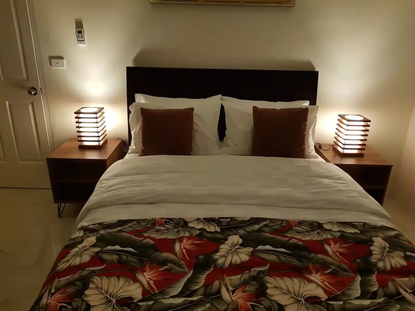 Una cama bien hecha con una colcha estampada, dos lámparas de noche y una mesita de noche con un libro.