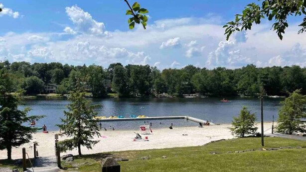 En park vid sjön med en sandstrand, ett område avsett för bad och en gräsyta med en träkonstruktion.