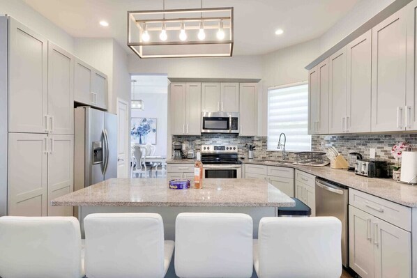 Una cocina moderna con gabinetes blancos, una isla central, electrodomésticos de acero inoxidable y un comedor con sillas blancas.