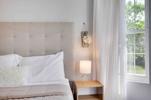 Un dormitorio con cama, una mesita de noche con lámpara, una ventana con cortinas y un aplique de pared.