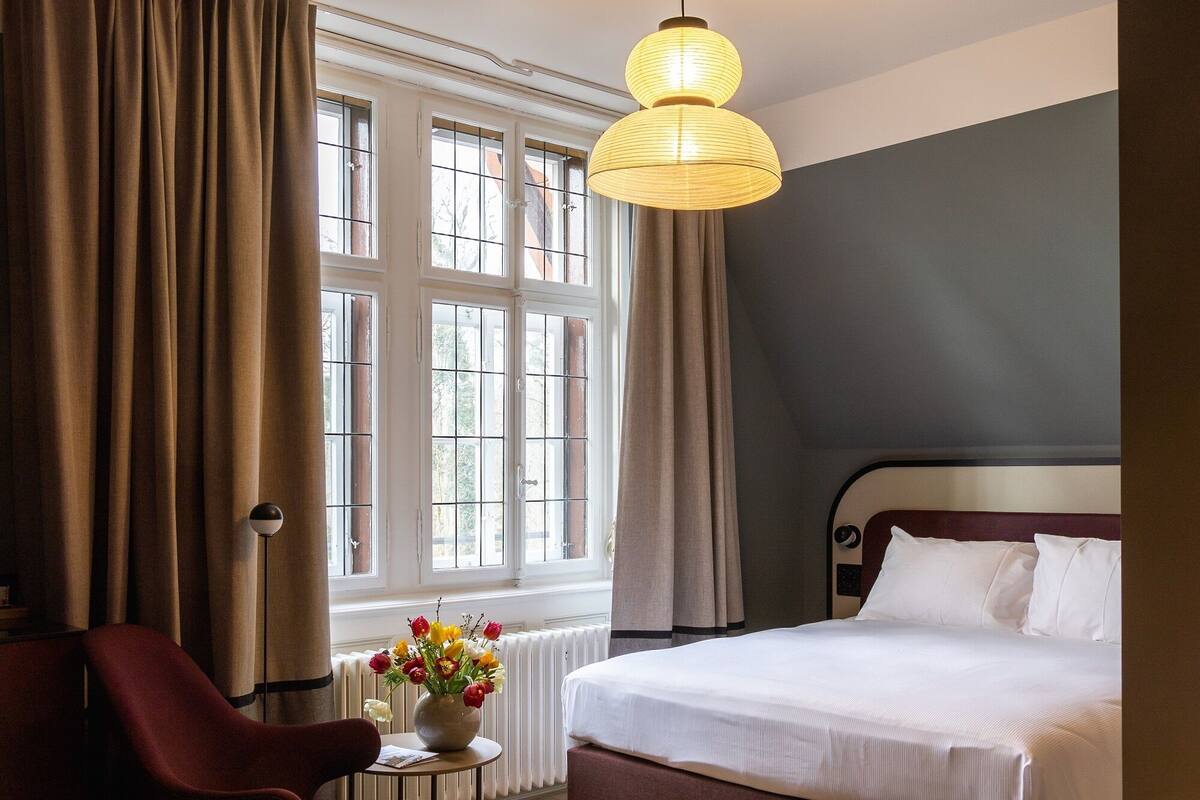 Una habitación de hotel con cama, una silla, una mesa con un jarrón de flores y ventanas grandes con cortinas.