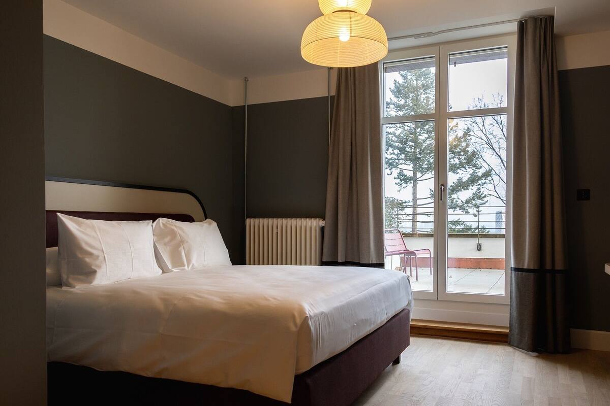 Un dormitorio con una cama grande, un radiador, una ventana con cortinas y vistas a árboles.