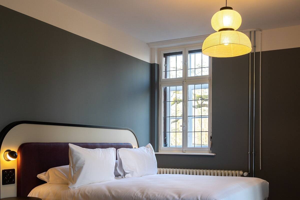 Un dormitorio con cama, ventana con persianas, radiador y lámpara colgante.