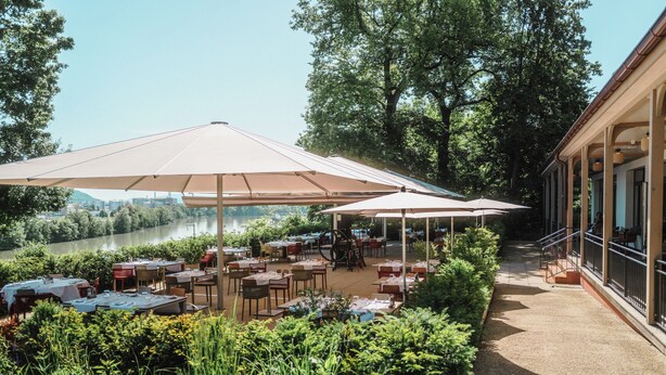 Un espacio para comer al aire libre con mesas y sombrillas, rodeado de follaje y vistas a un río.