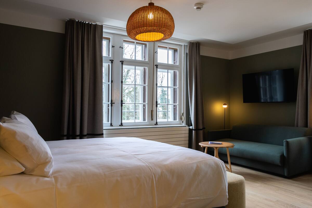 Una habitación de hotel con una cama grande, un sofá, un televisor y una ventana con cortinas.