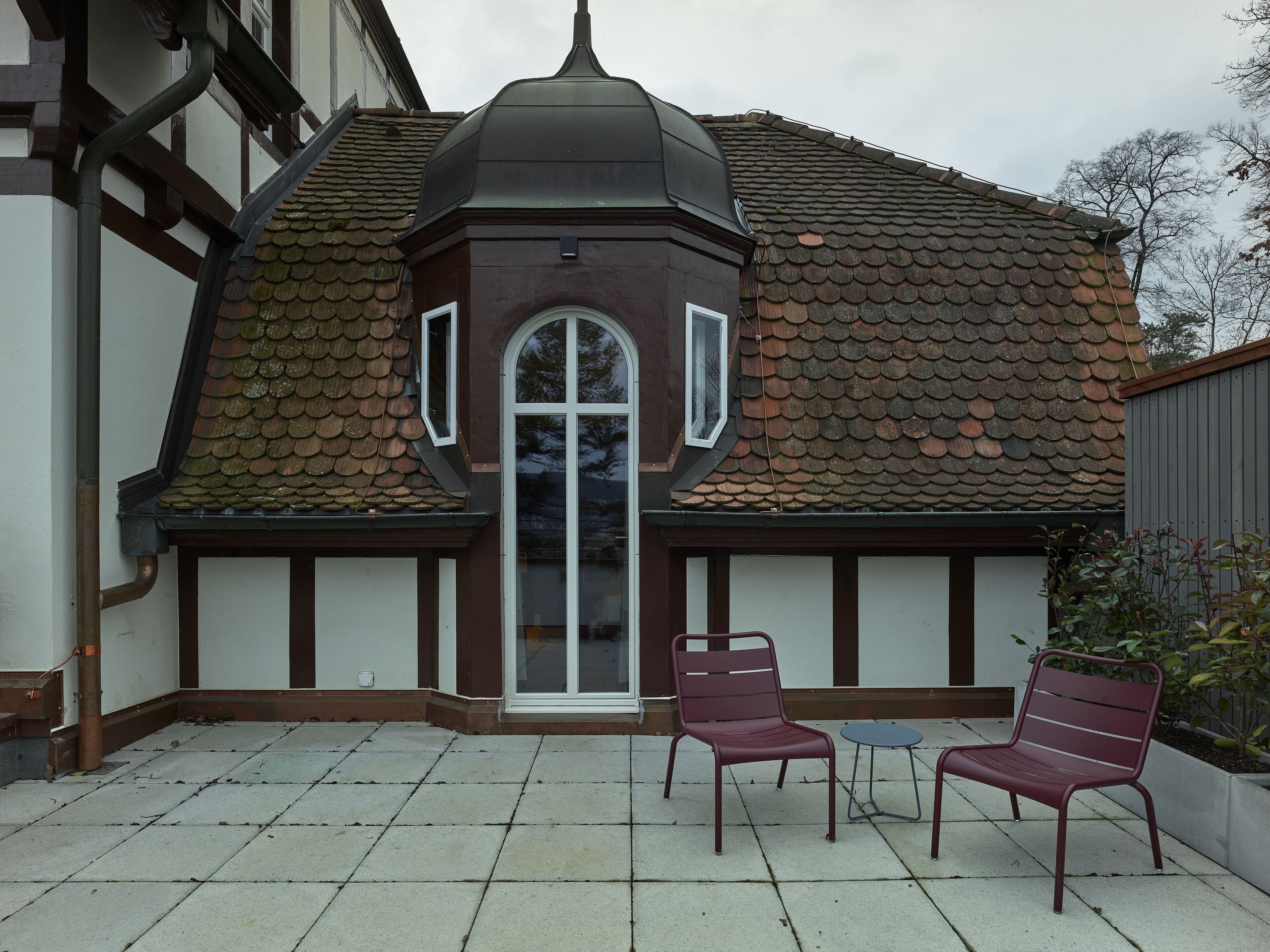Un bâtiment doté d’un toit recouvert de tuiles, d’une fenêtre centrale en arc de cercle et de deux chaises en métal accompagnées d’une petite table.