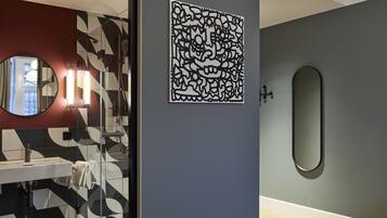 Un baño moderno con cabina de ducha de vidrio, un espejo redondo y una obra de arte geométrico en la pared.