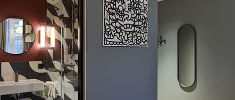 Un baño moderno con cabina de ducha de vidrio, un espejo redondo y una obra de arte geométrico en la pared.