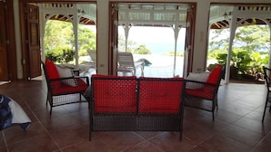 A room with a view of the ocean, featuring wicker furniture with red cushions, tiled flooring, and wooden ceiling beams. - Beautiful, secluded, open plan cottage with spectacular sea views (Castara)