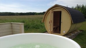 Een hottub met een houten terras ervoor, een houten sauna en een grasveld op de achtergrond.