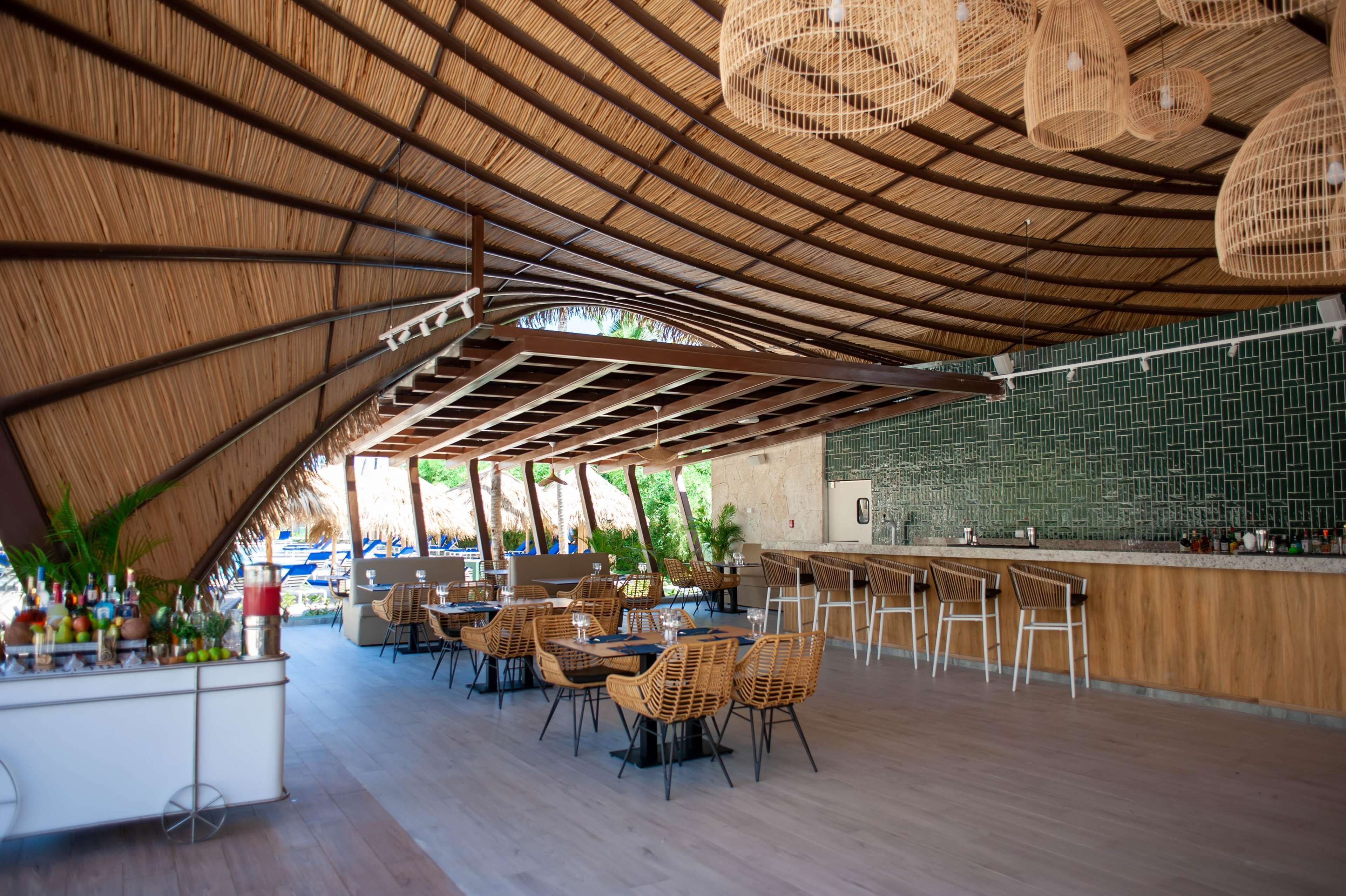 Ein Essbereich im Freien mit einem Reetdach, Korbmöbeln und einer Bar mit dunkelgrünen Fliesen.