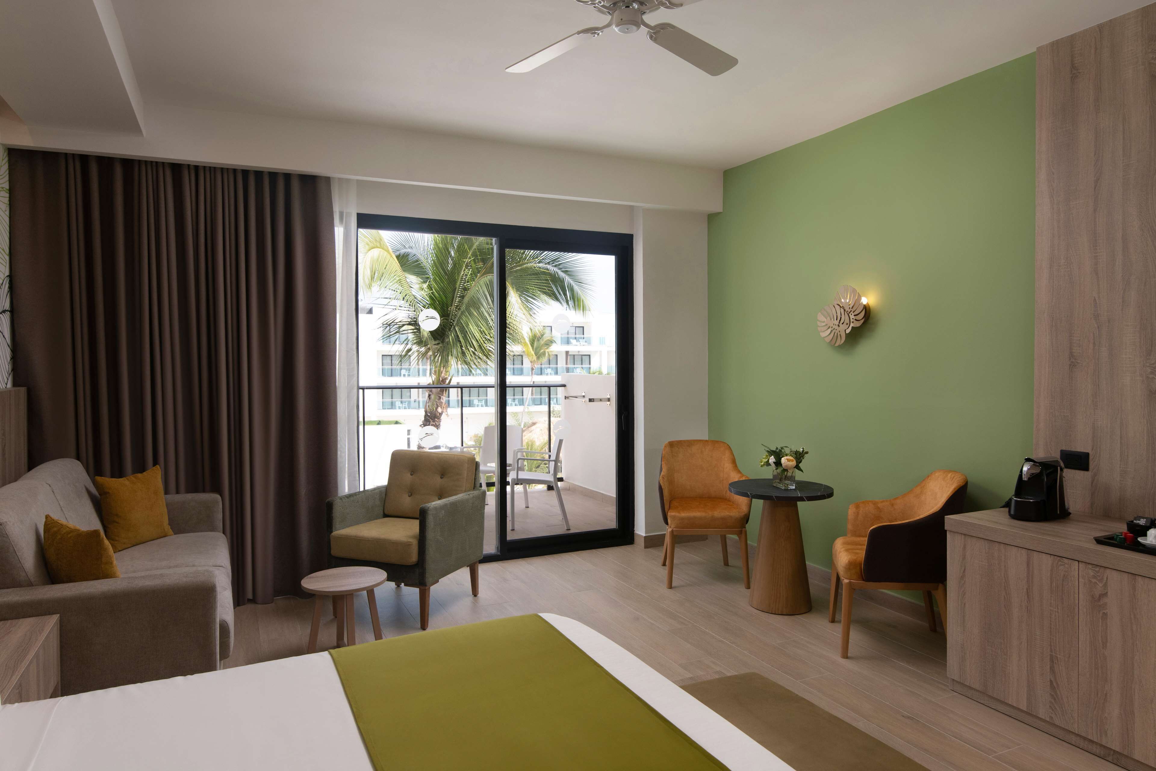 Une chambre d’hôtel comprenant un lit, un canapé, deux fauteuils, une petite table, une armoire et un balcon avec vue sur les palmiers et les immeubles.