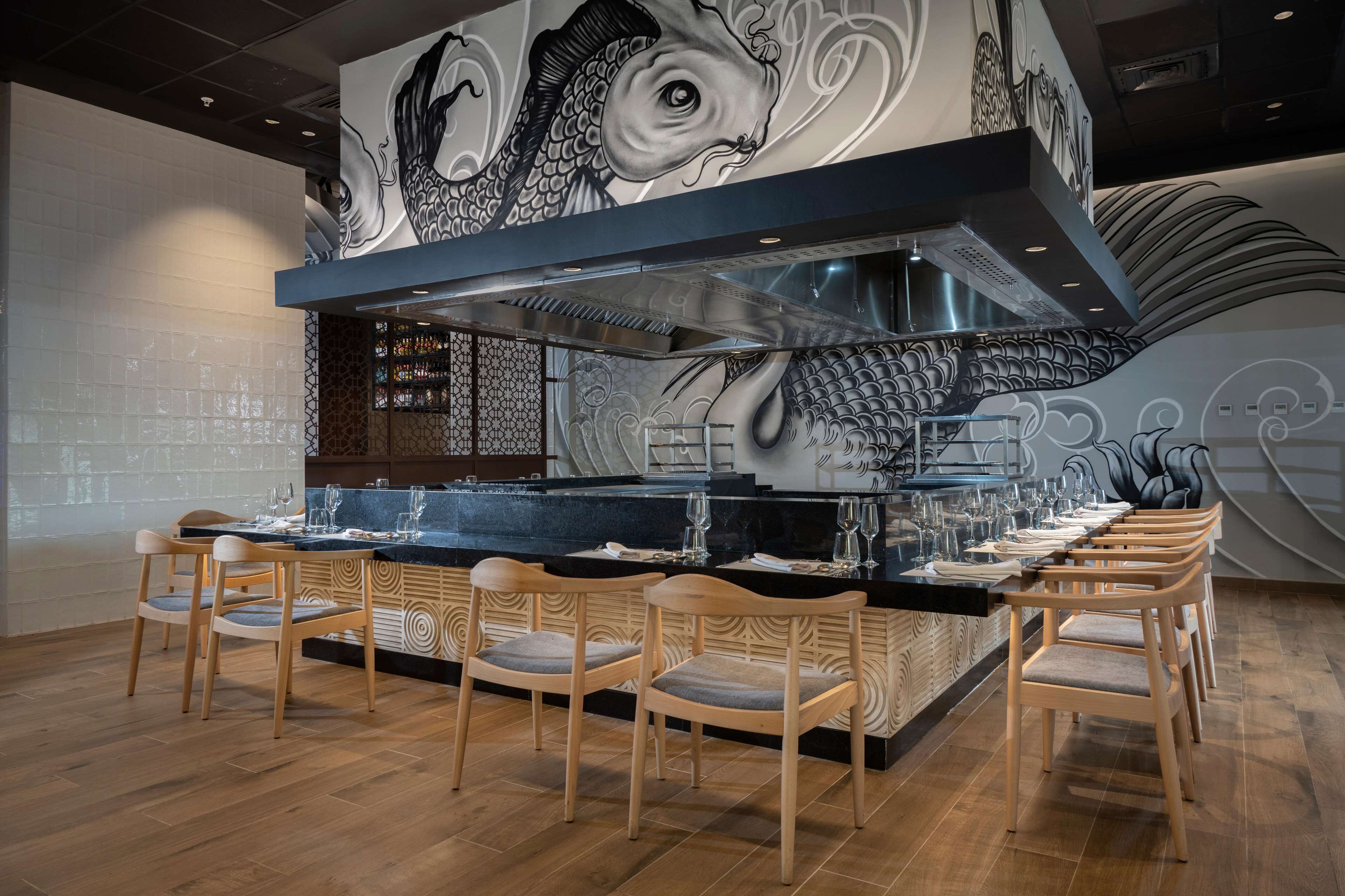 Uma área de bar moderna com um balcão comprido, cadeiras de madeira e um mural decorativo com peixes na parede.