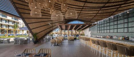 A spacious outdoor dining area with wicker chairs and tables, a bar with stools, and a thatched roof ceiling.