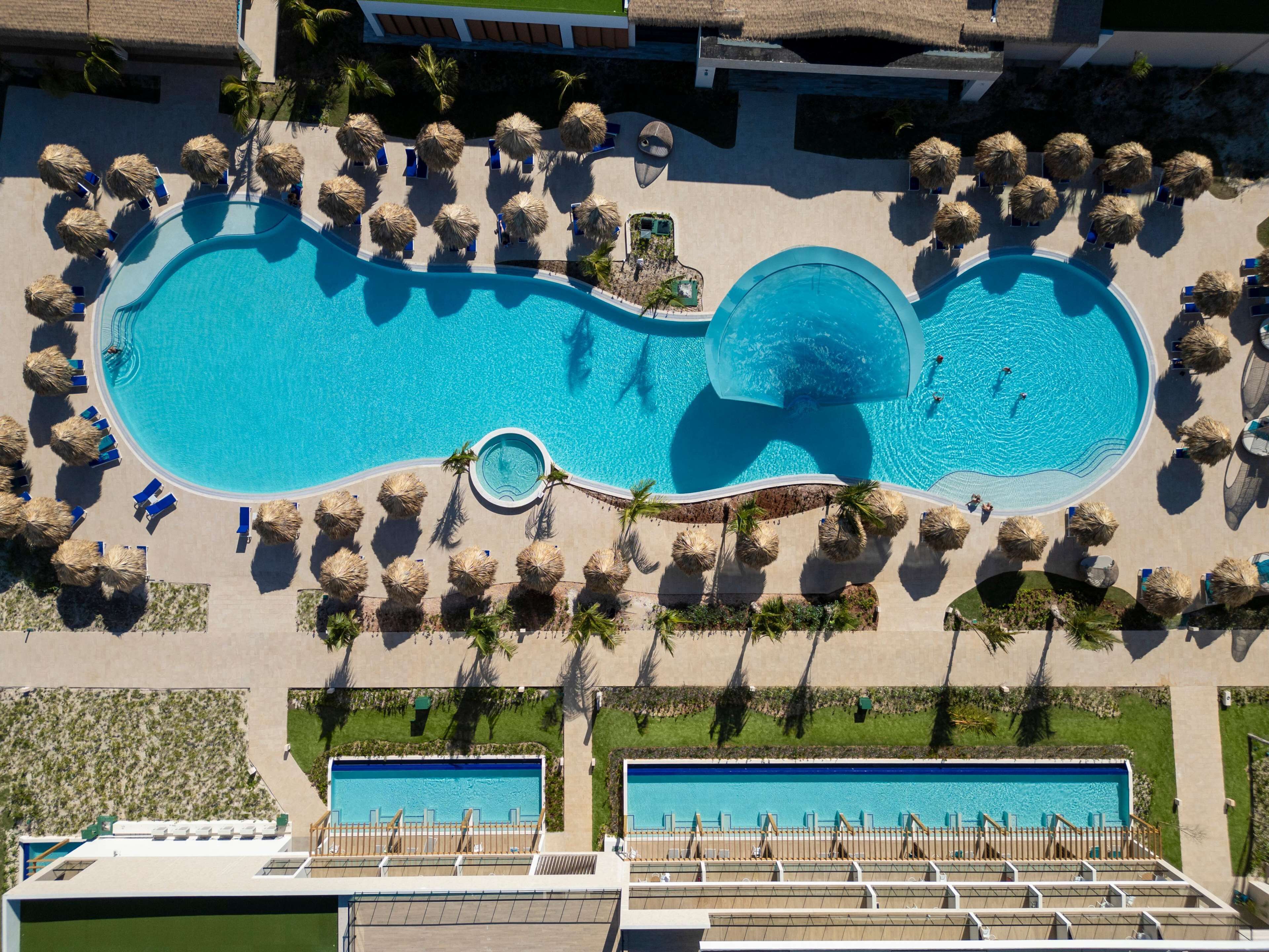 Vista aérea de un resort con una amplia piscina central rodeada de tumbonas y palmeras.