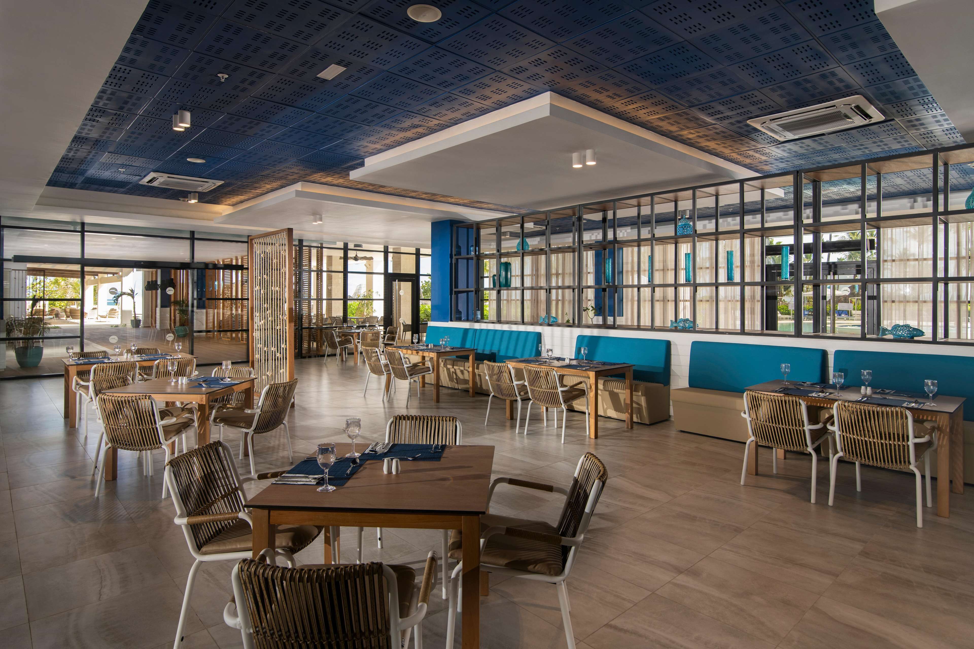 En rummelig spiseplads med flere borde og stole, store vinduer og et moderne loftdesign.
