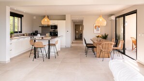 Una cocina moderna con armarios blancos, una isla central y taburetes de bar. Hay un comedor con mesa de madera y sillas de mimbre.