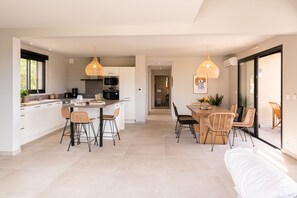 A modern kitchen with white cabinets, a central island, and bar stools. There is a dining area with a wooden table and wicker chairs. - Villa 1: Cap Camarat (Roquebrune-sur-Argens)