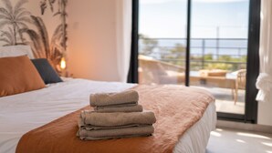 Habitación de hotel con cama, una pila de toallas, un balcón con vistas al mar y un muro cubierto de plantas tropicales.