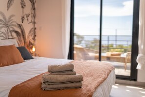 A hotel room with a bed, a stack of towels, a balcony with a view of the sea, and a tropical plant wall. - Villa 1: Cap Camarat (Roquebrune-sur-Argens)