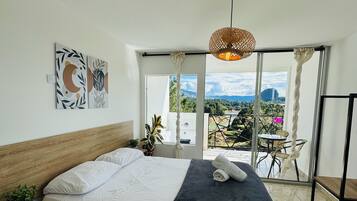 Un dormitorio con cama, una mesita de noche con una planta, vistas al exterior a través de un ventanal grande, y una lámpara colgante.