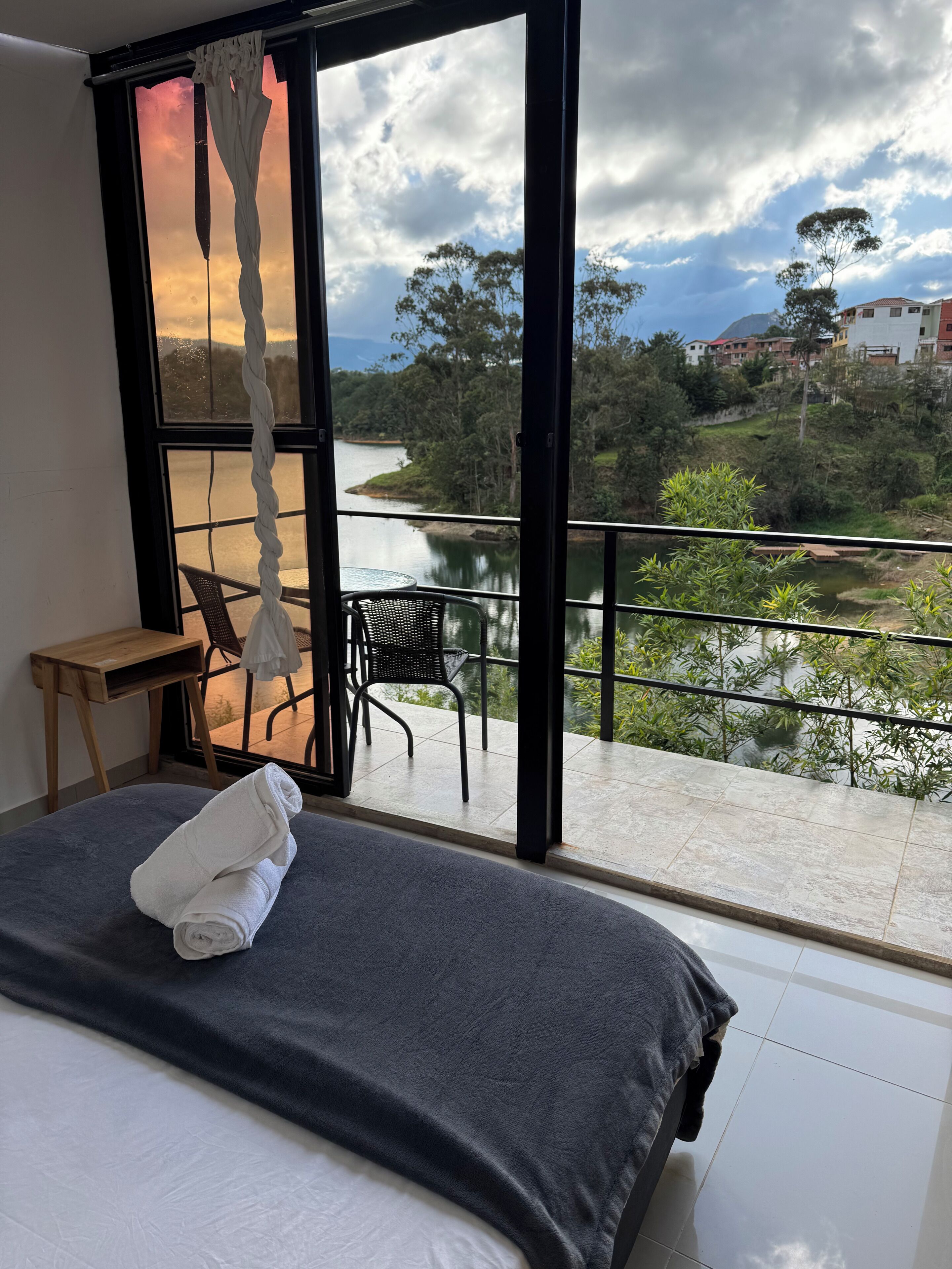 A bedroom with a bed, a chair, and a table. The room has a large window with a view of a river and greenery.