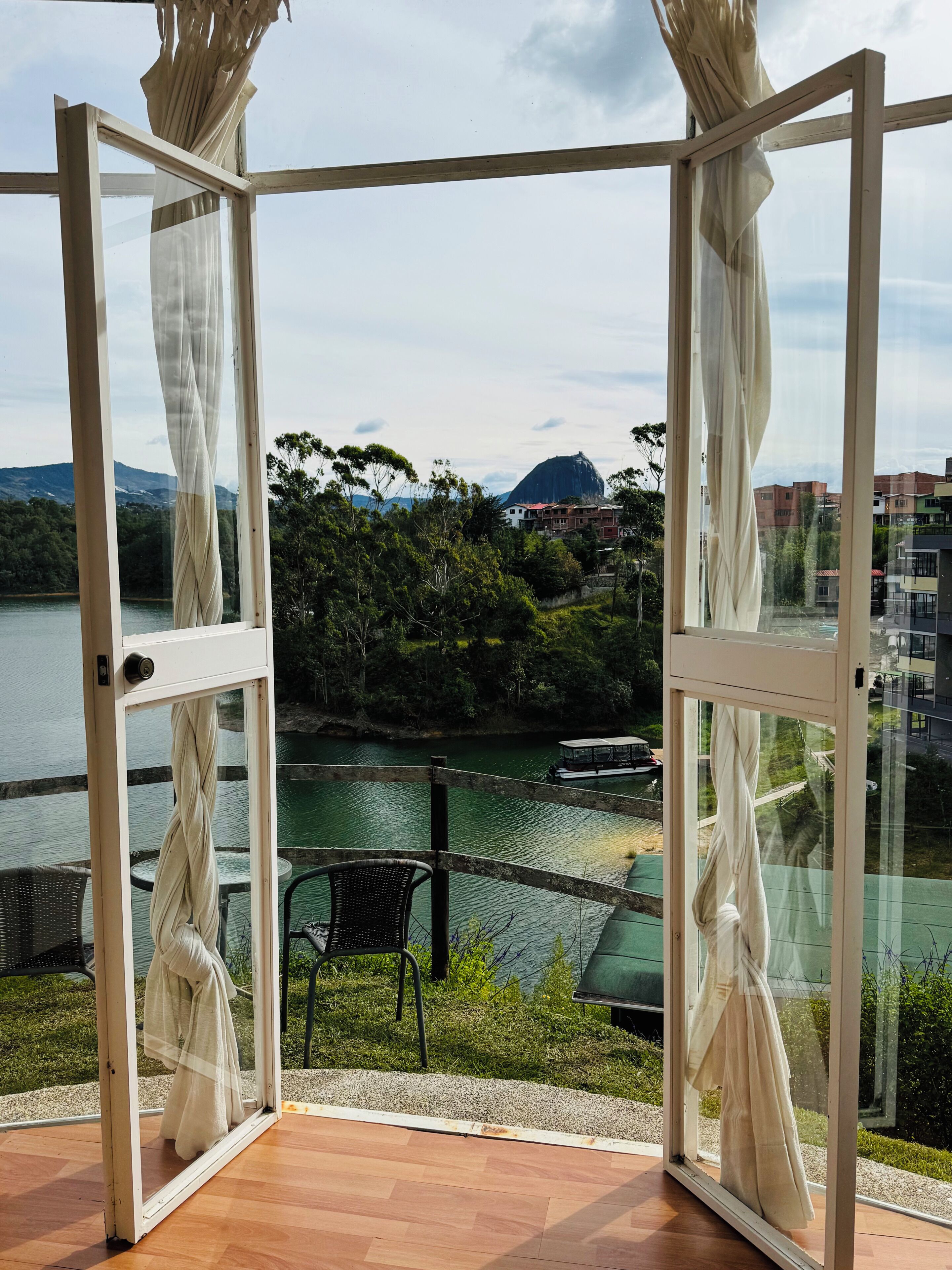 An open glass door leading to a balcony with a view of a lake, greenery, and buildings in the distance.