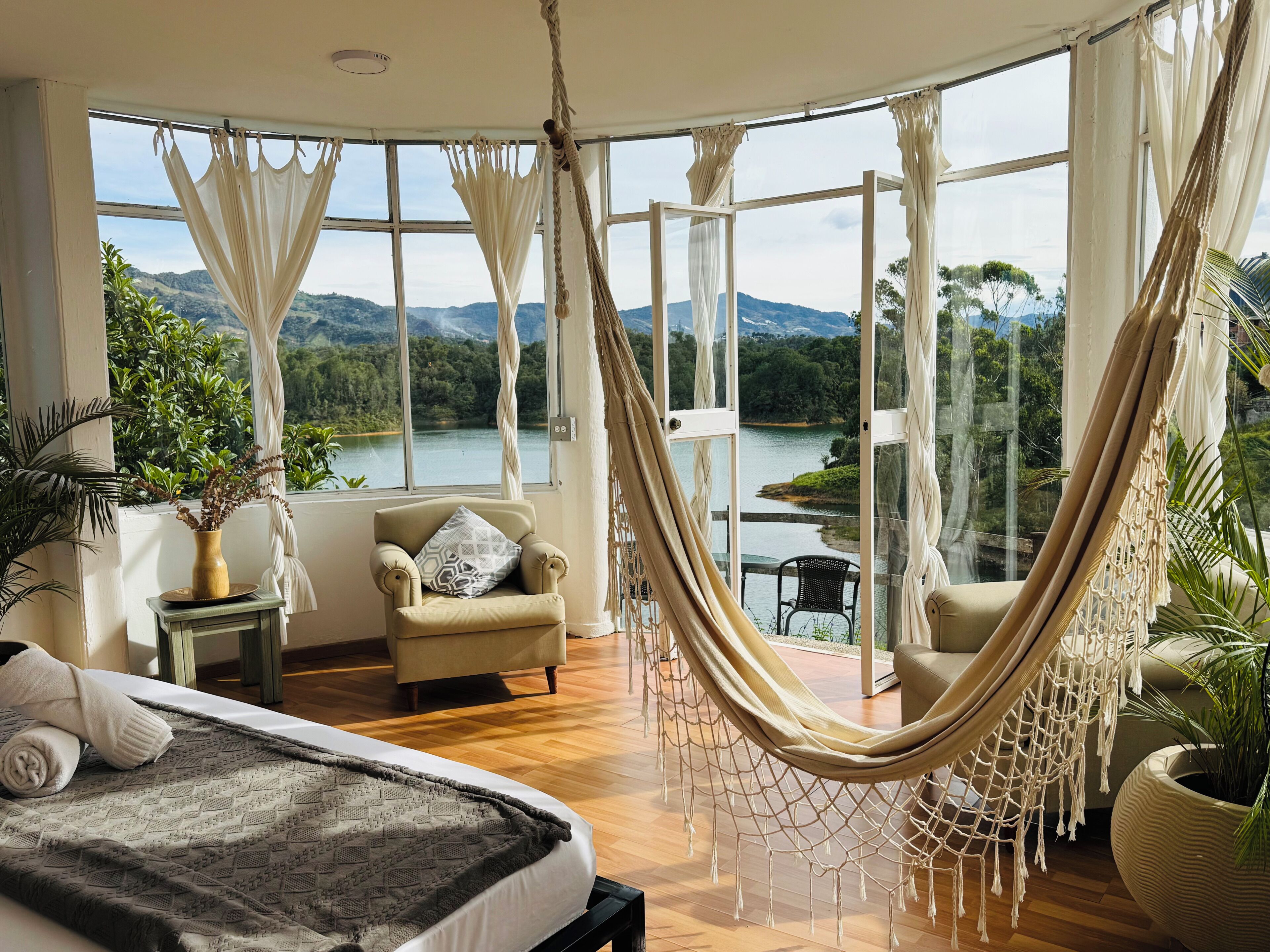 Un dormitorio con un ventanal grande, una hamaca, una cama, una silla y vistas a un lago y montañas.