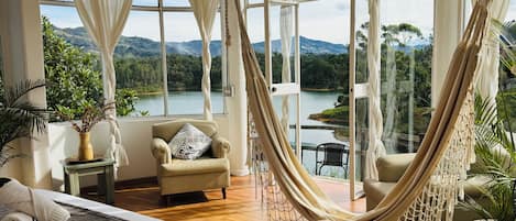 Un dormitorio con un ventanal grande, una hamaca, una cama, una silla y vistas a un lago y montañas.
