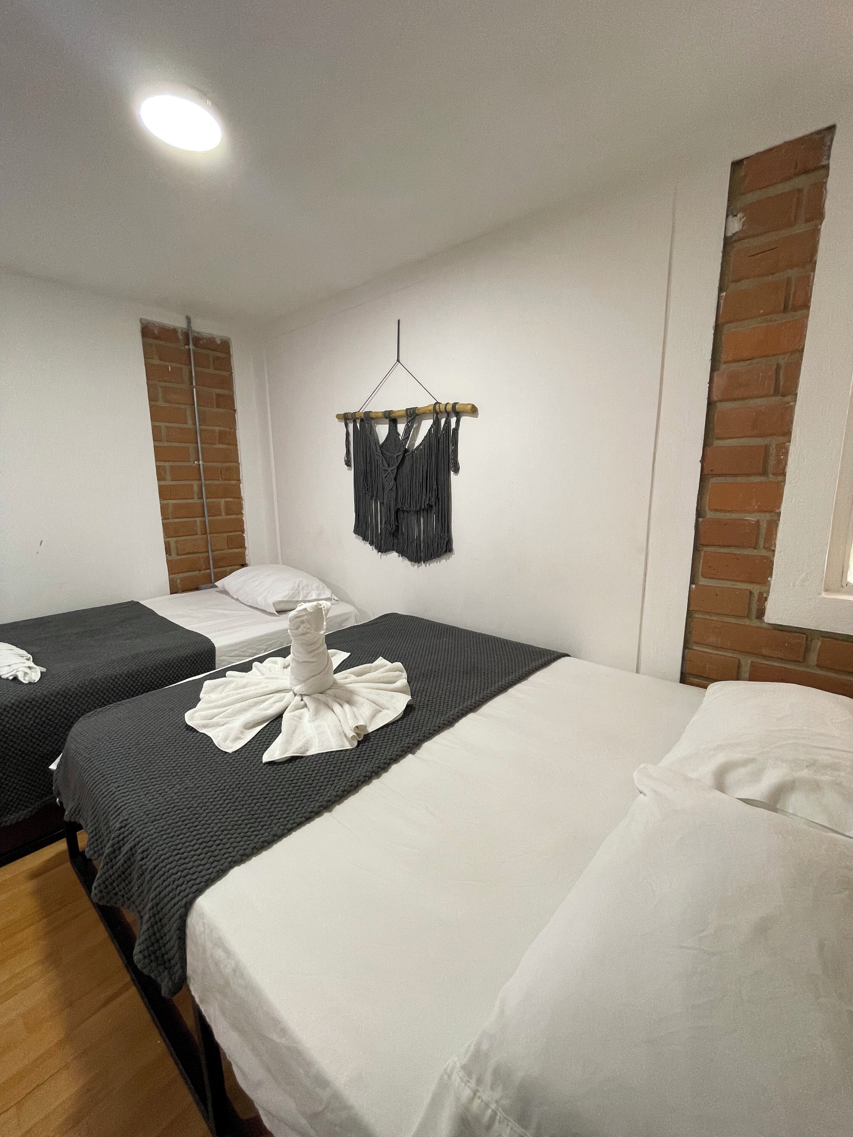 Quarto de hotel com duas camas, uma decoração de macramé pendurada na parede e uma parede de tijolos.