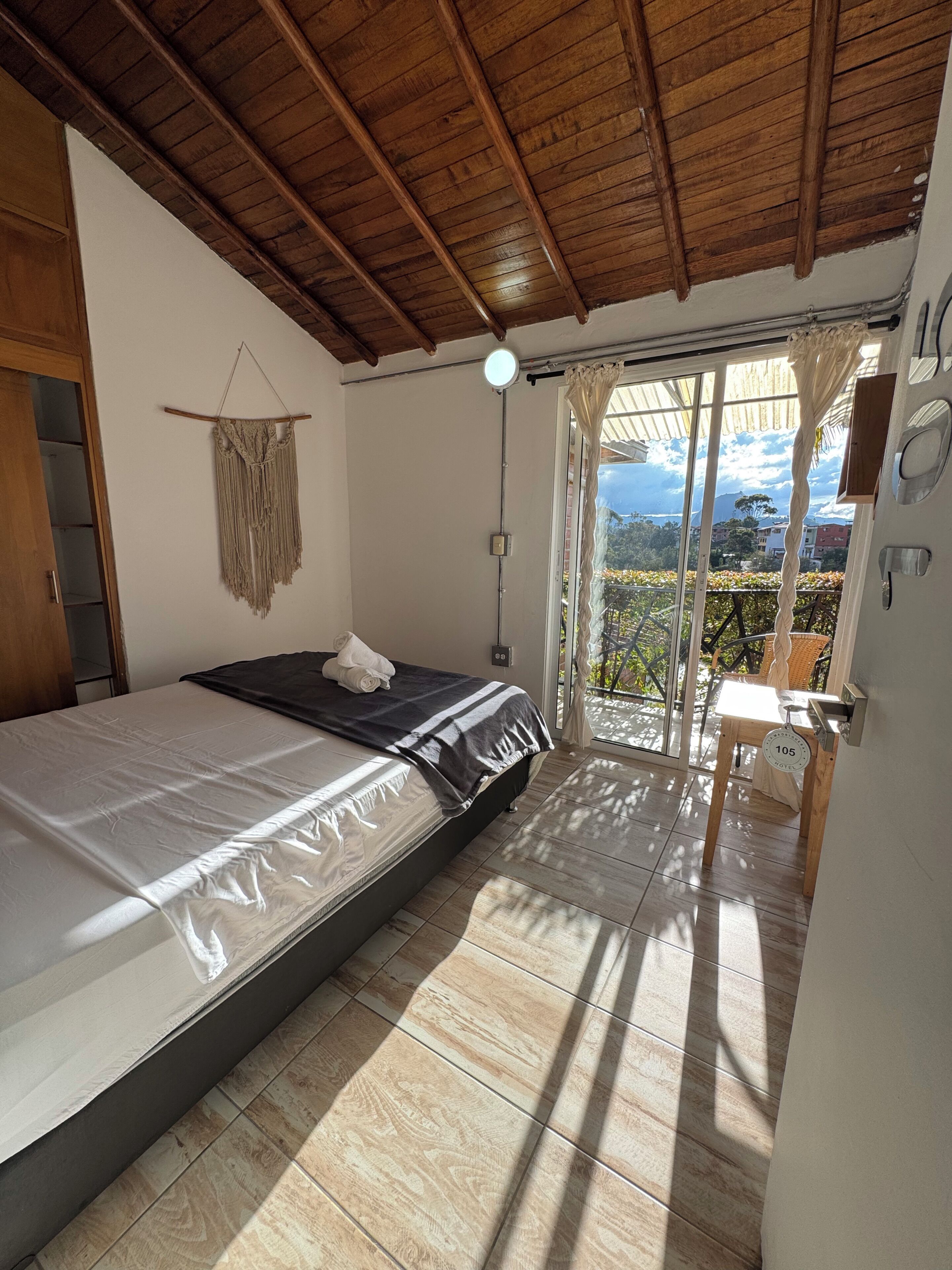 Een slaapkamer met een bed, houten plafond, raam met gordijnen en uitzicht op buiten.