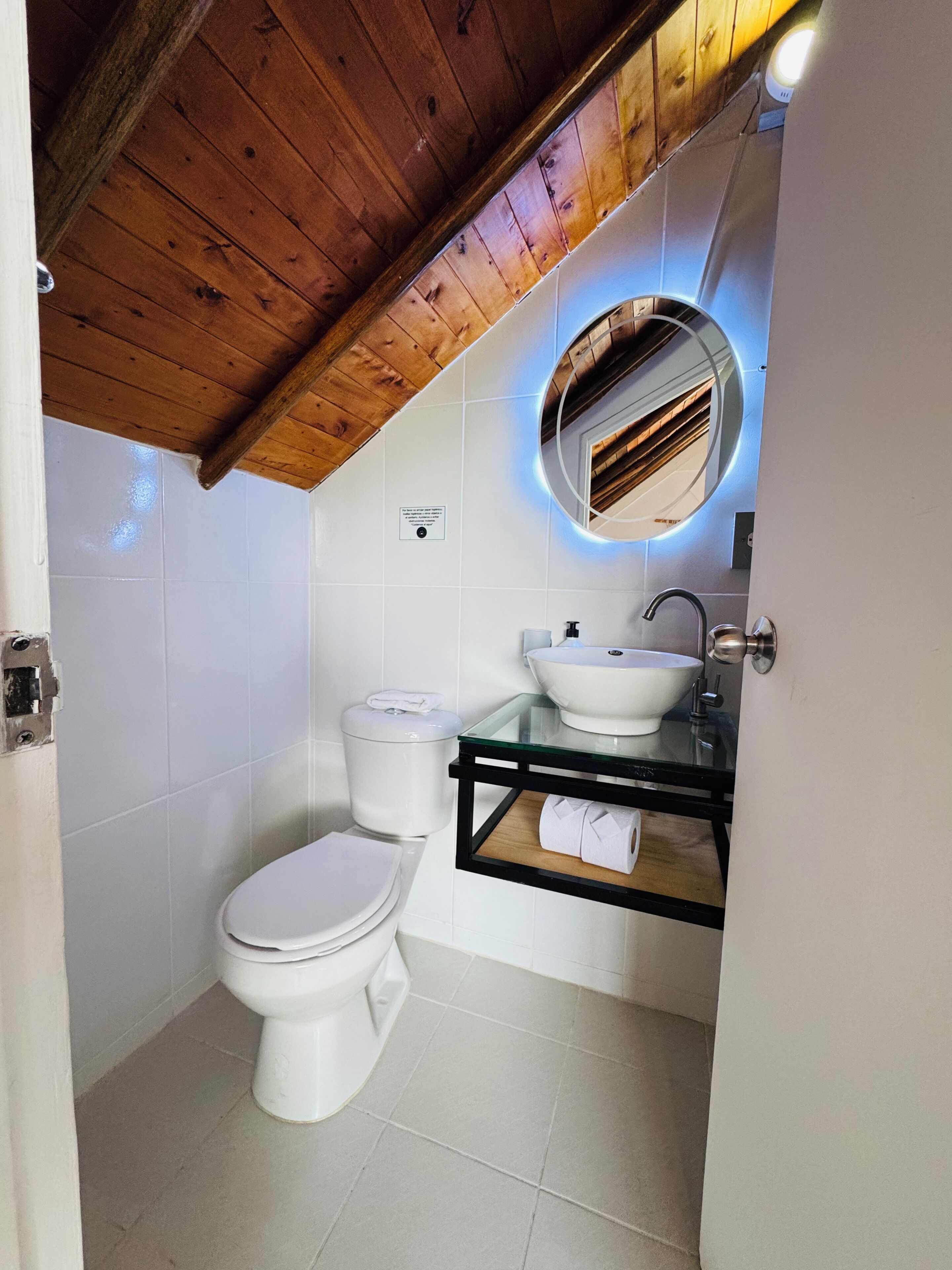 Un baño con techo de madera, inodoro, lavamanos con espejo y estante de vidrio.