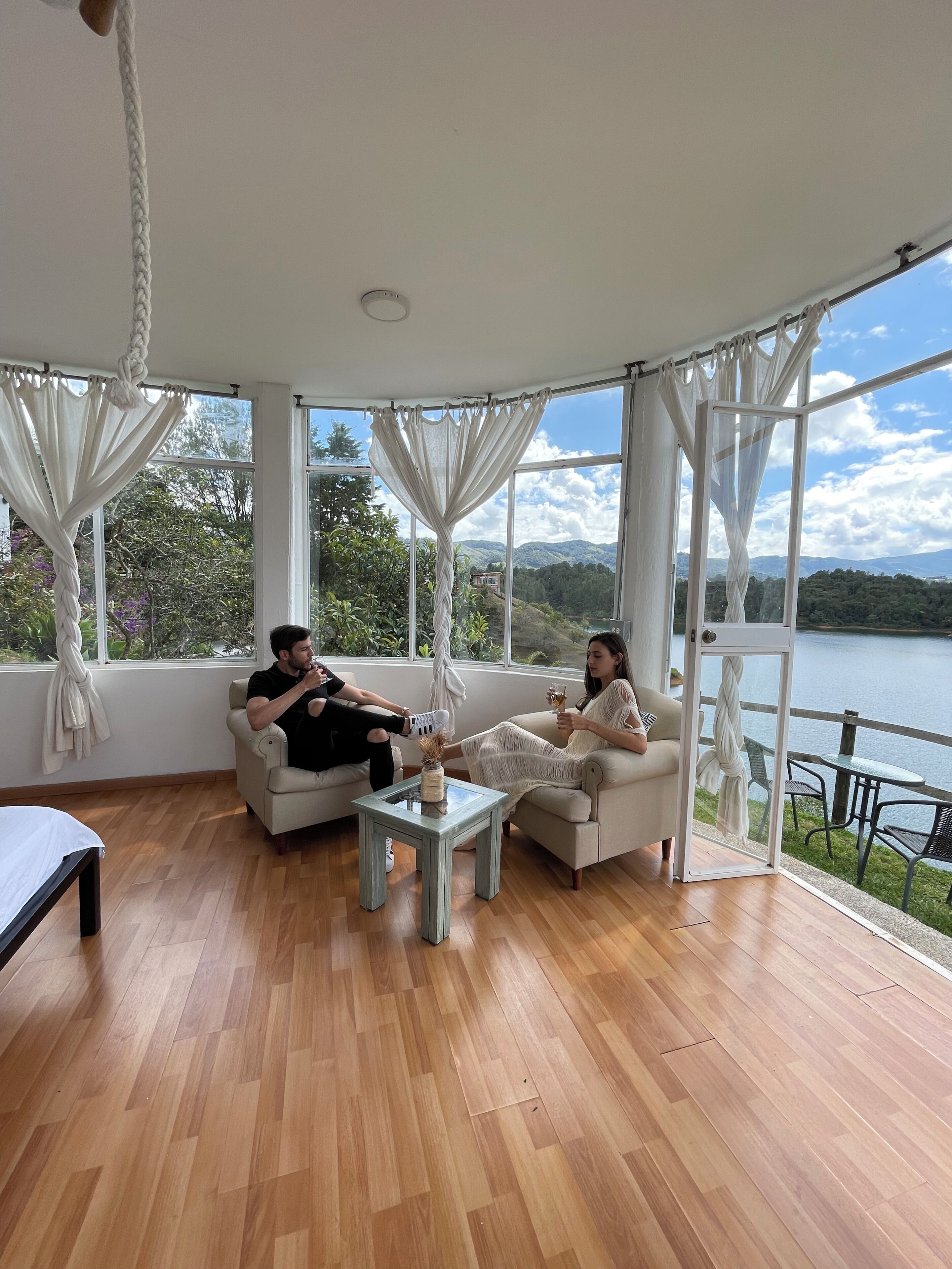 A room with a wooden floor, two white sofas, a small glass table, and a view of a lake and mountains through large windows.