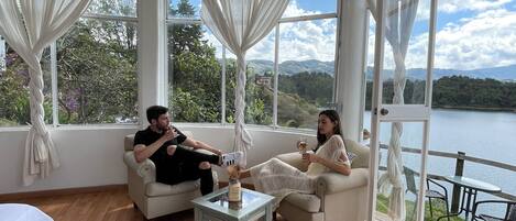 A room with a wooden floor, two white sofas, a small glass table, and a view of a lake and mountains through large windows.