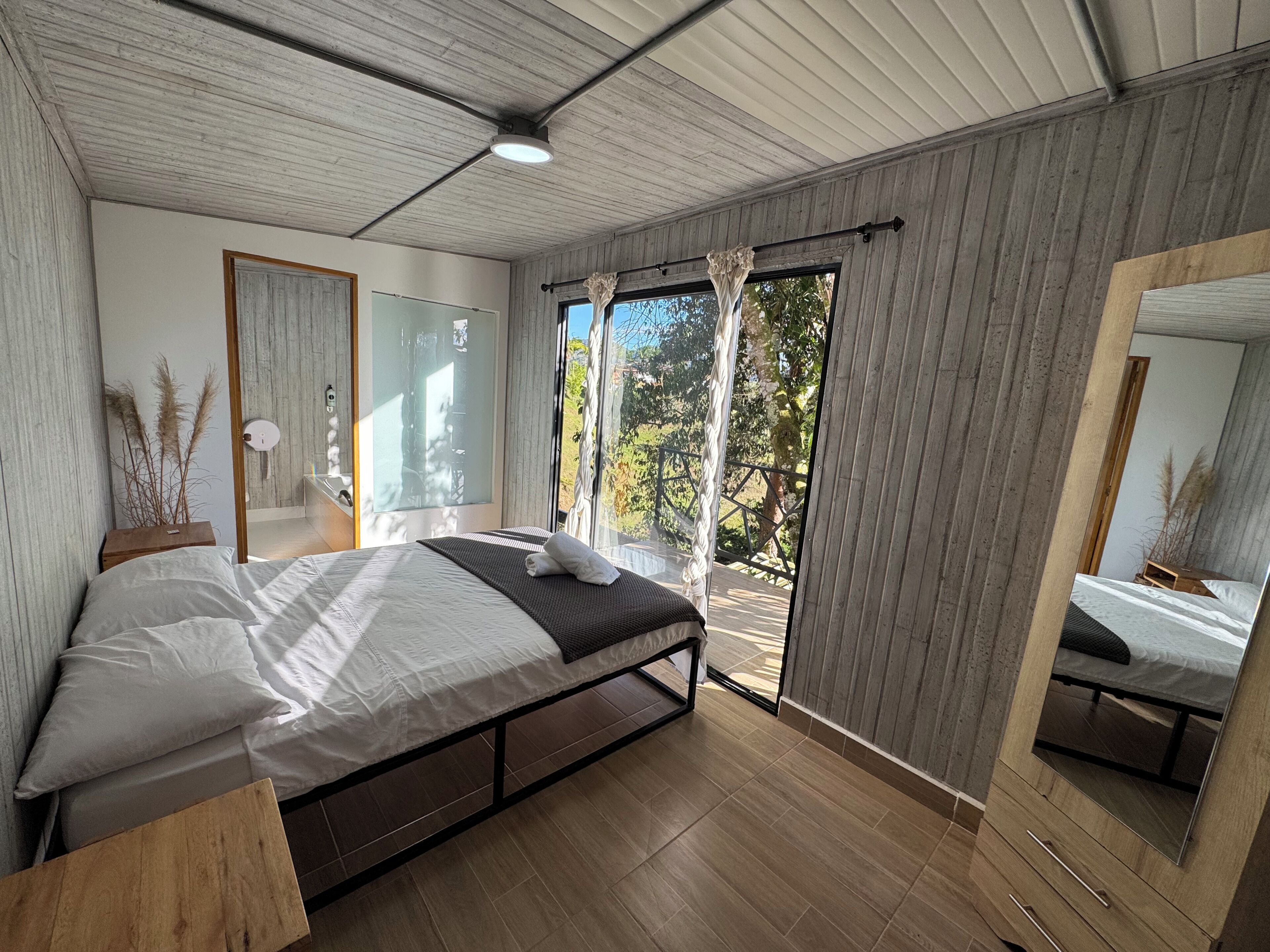Een slaapkamer met een bed, houten vloer, groot raam en spiegel.