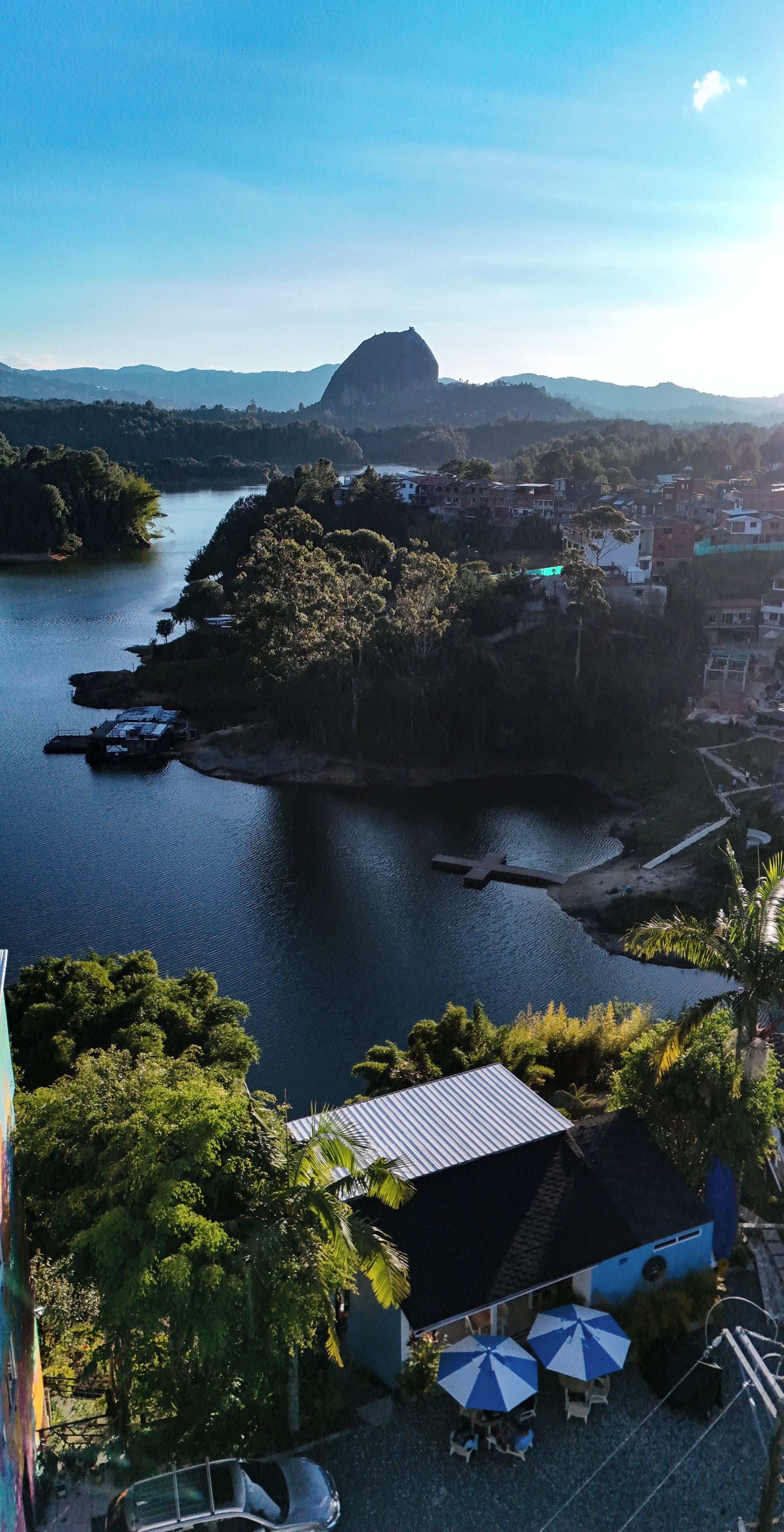 Paisagem costeira com uma formação rochosa imponente, um corpo d'água e um pequeno povoado com edifícios e vegetação.