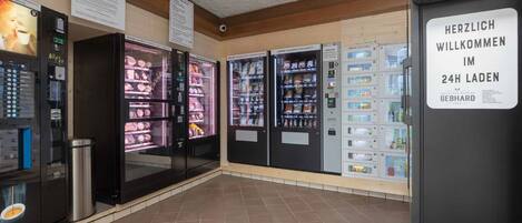 Zona de venta automática abierta las 24 horas con diversas opciones de comida y bebida, incluyendo productos refrigerados y una máquina de café.