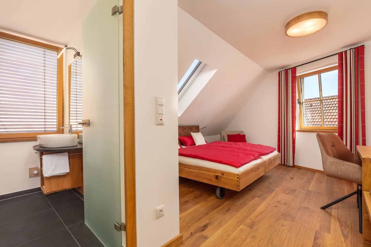 Une chambre à coucher avec un lit en bois, une couverture rouge, une chaise et une fenêtre avec des rideaux.
