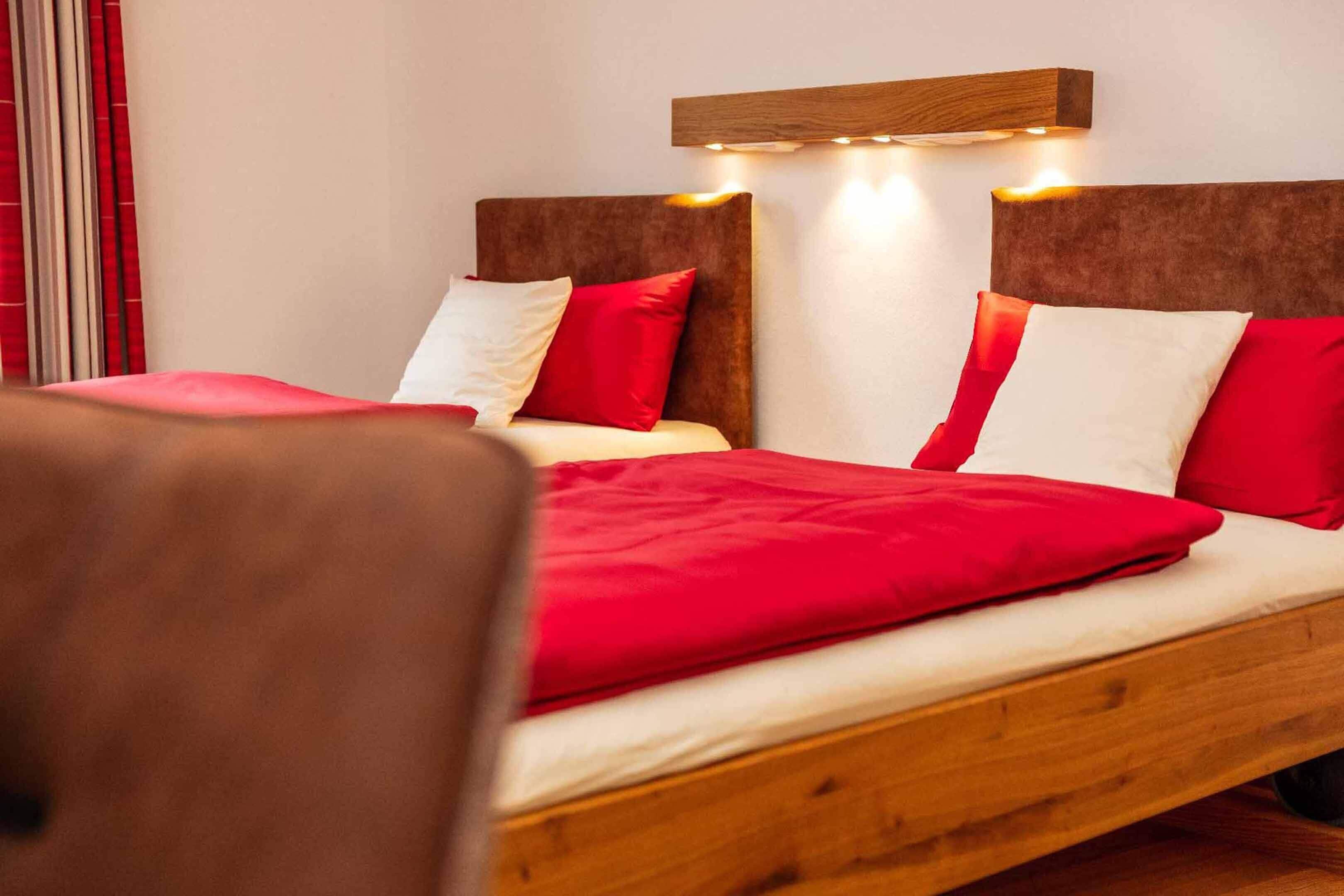 Ein Doppelbett mit Holzrahmen, Bettwäsche in Rot und Weiß sowie ein Kopfteil mit integrierter Beleuchtung.
