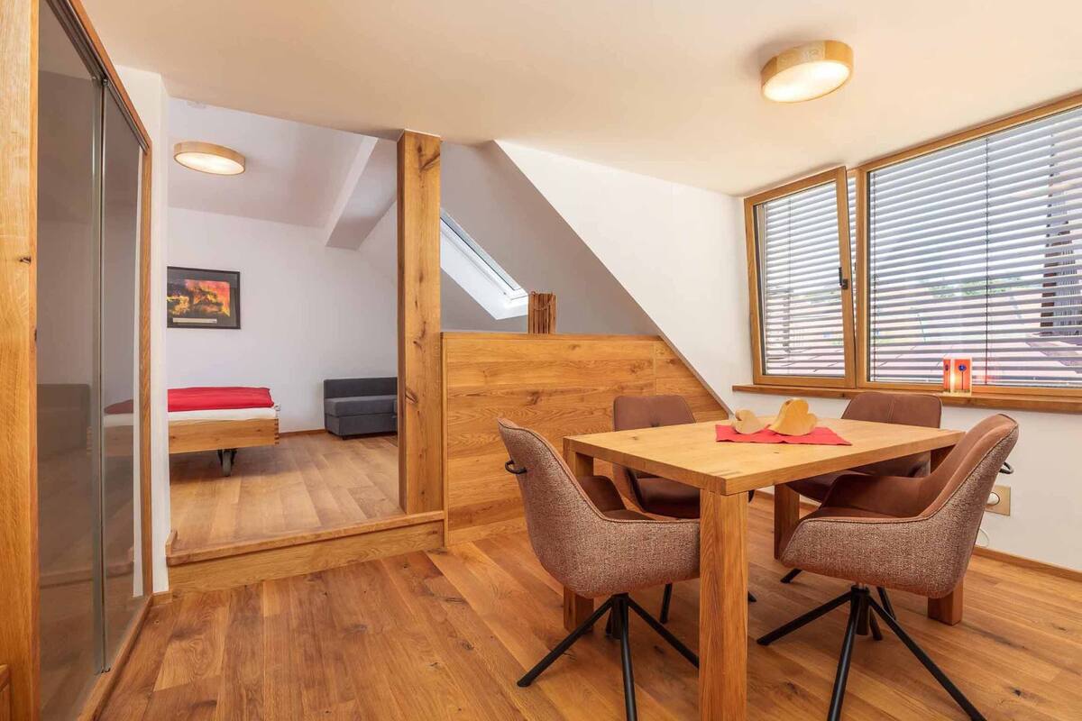Une pièce de taille modeste, comprenant une table à manger en bois et quatre chaises, un petit lit avec un matelas rouge, un canapé gris et une cloison en bois.