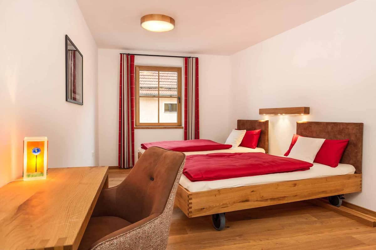 Une chambre avec deux lits, un bureau en bois, une chaise, une fenêtre avec des rideaux rouges et un luminaire fixé au mur.