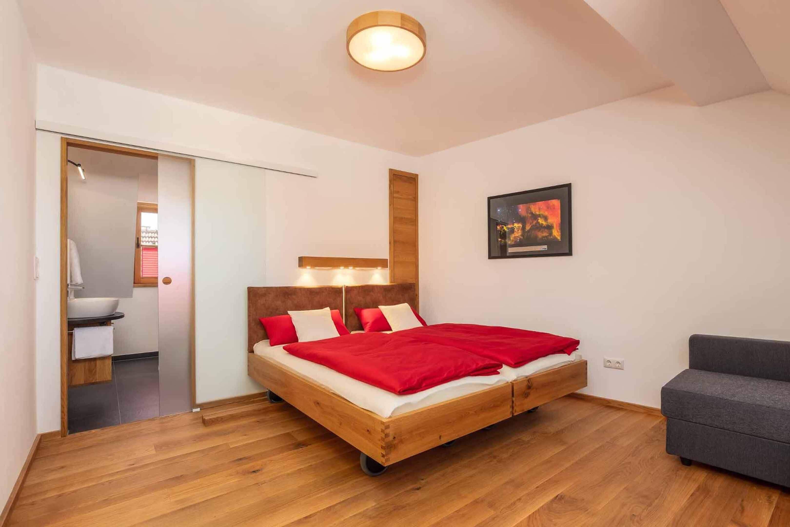 A bedroom with a wooden bed, a gray sofa, a wooden headboard, a wall-mounted light, and a framed picture.
