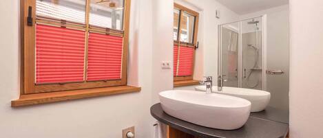A modern bathroom with a large mirror, two sinks, a shower, and a window with blinds.