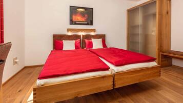 A modern bedroom with a wooden bed, red bedding, a wall-mounted artwork, and a wooden wardrobe.
