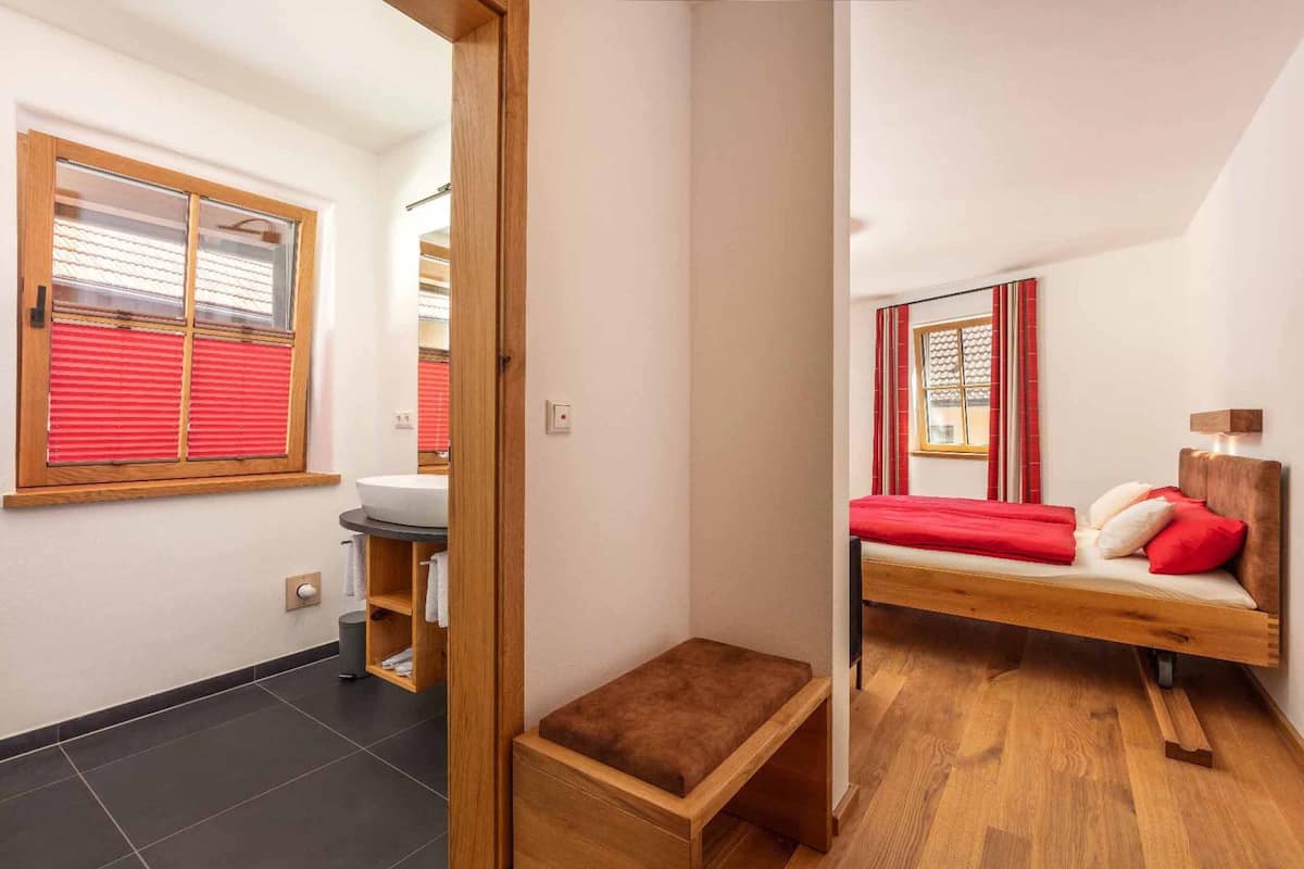 Une chambre à coucher de taille modeste, avec un lit en bois, une petite table de chevet, un rideau rouge et une fenêtre avec des stores rouges.