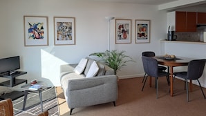 A modern living room with a grey sofa, a coffee table, a TV, and a dining area with a table and chairs. There are three framed artworks on the wall.