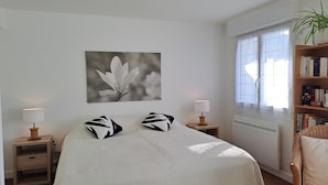 A bedroom with a bed, two nightstands, a bedside lamp, a radiator, a window with curtains, a bookshelf, and a wall-mounted picture.